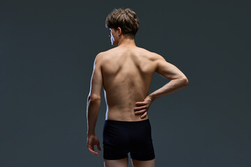 Fototapeta na wymiar Rear view portrait of young brunette man posing, in underwear holding his muscular, healthy back isolated over dark grey studio background.
