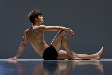 Full-length photo of young man with muscular build body posing in underwear sitting on floor over dark grey studio background.