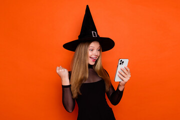 Photo of lucky excited lady wear black wizard costume cap winning halloween game modern gadget isolated orange color background