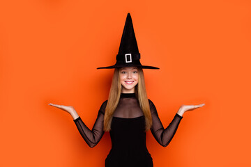 Portrait of charming teen schoolgirl wizard costume arms hold empty space vs scales isolated on orange color background