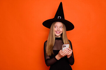 Photo portrait of pretty blonde teenager girl device look empty space dressed black halloween outfit isolated on orange color background