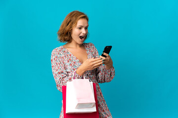 Young Georgian woman with shopping bag isolated on blue background holding shopping bags and writing a message with her cell phone to a friend