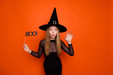 Photo portrait of pretty blonde teenager girl hold boo sign scratch hand dressed black halloween outfit isolated on orange color background