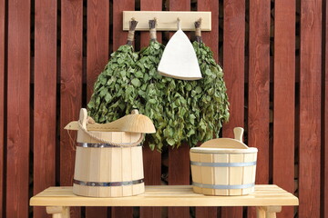 A wooden sauna accessories are on a bench standing in front of red board wall of firewood shed with hanging dry birch brooms on it. Outdoors. 