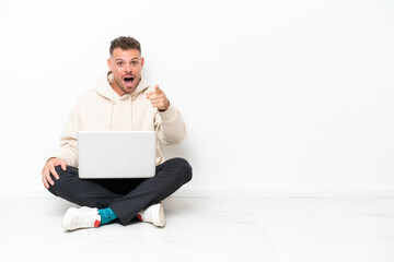 Young caucasian man with a laptop sitting on the floor isolated on white background surprised and pointing front