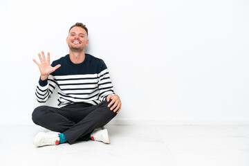 Young man sitting on the floor isolated on white background counting five with fingers