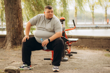 One tired young overweight man sitting down at park bench to rest. A fat person resting outdoors