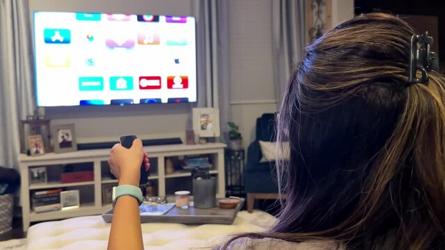 Rear view of woman scrolling streaming apps on TV, using remote control, at home