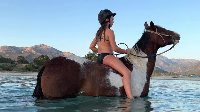 Young red-haired girl in bikini have fun bathing and bareback riding horse in sea water in summer season