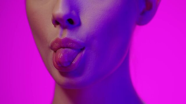 Close up of a young pretty woman with beautiful red lips while sticking out her tongue with purple light in her face against purple background in slow motion