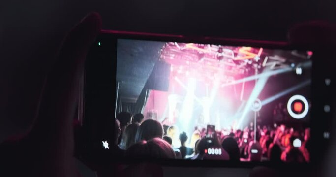 Closeup, phone screen filming band concert. Girl holding phone camera in hands at rock band concert. Pink magenta bright spotlights flashing in stage. Audience at festival event. Disco entertainment