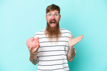 Redhead man with long beard holding a piggybank isolated on blue background with shocked facial...