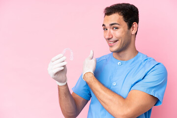 Dentist caucasian man holding invisible braces isolated on pink background pointing back