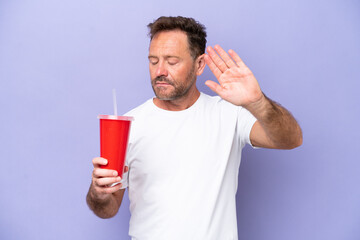 Middle age caucasian man holding soda isolated on purple background making stop gesture and disappointed