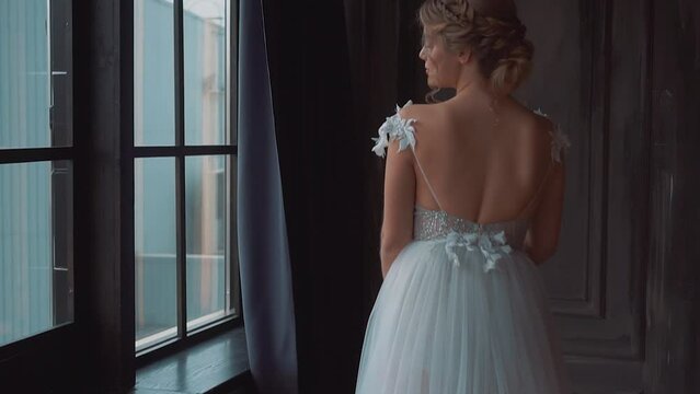 Luxurious young blonde woman stands at window waiting love, dark classic interior room. Bride girl in white wedding day dress, bare back open. Professional hairstyle, fantasy fairy princess style 