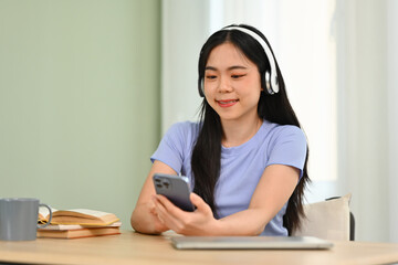 Smiling young asian girl wearing headphone and using smartphone at desk in living room