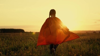 Little girl with long red cloak walks across sunset meadow superhero party