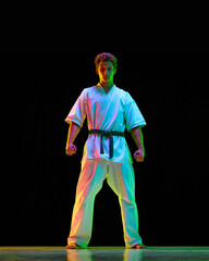 Full-length portrait of young sportive guy, karate athlete in white kimono standing over black studio background in neon lights. Concept of martial arts, combat sport, energy, strength, health. Ad