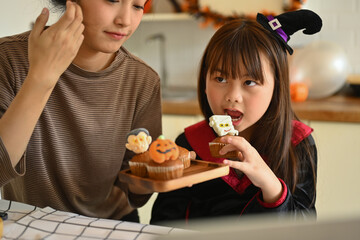 Cute little asian girl dressed as a witch eating colorful Halloween cupcakes with mother in kitchen. People, holiday and festival