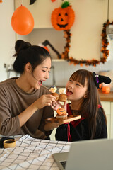 Cute little asian girl dressed as a witch eating colorful Halloween cupcakes with mother in kitchen. People, holiday and festival