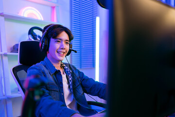 A young Asian man professional gamer wearing jeans jacket sits on a chair with a gaming table with pc, keyboard, monitor, microphone. Prepare for competition, cast gameplay or record a podcast.