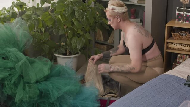 Confident Caucasian queer man wearing bra and nide tights taking blond wig from suitcase with fancy clothes and accessories while getting dressed for drag show at homeConfident Caucasian queer man wea