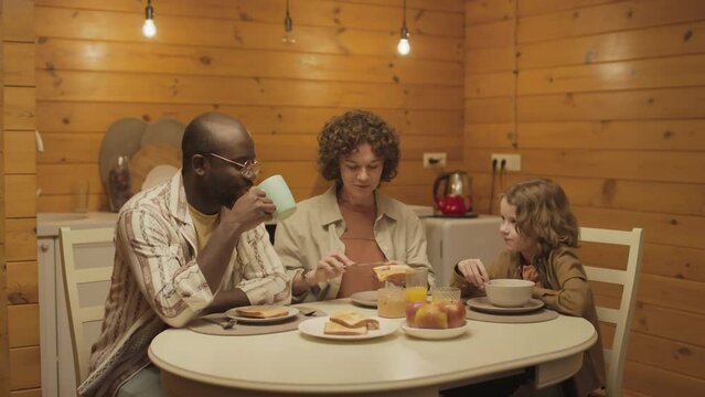 Medium shot of ethnically diverse family of three sitting around table having meal together and talking with each other