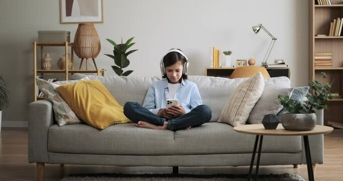 Young boy wearing headphones, immerses himself in the world of music while browsing content on phone. Seated comfortably on the living room couch, he finds solace in the melodies.