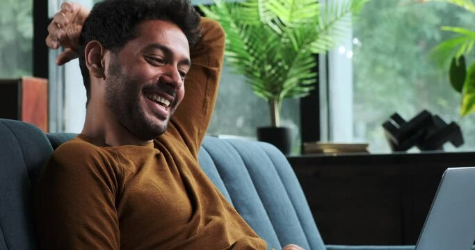Middle Eastern man sits on a sofa, using laptop and sharing a moment of laughter. His relaxed and cheerful demeanor exemplifies the balance of work and leisure in a comfortable home setting.