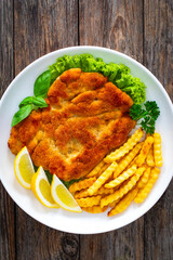 Crispy breaded seared chicken cutlet with French fries,   lemon and fresh vegetables on wooden table
