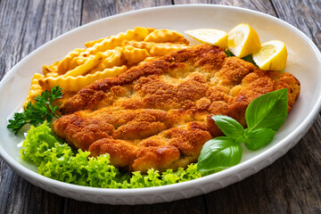 Crispy breaded seared chicken cutlet with French fries,   lemon and fresh vegetables on wooden table
