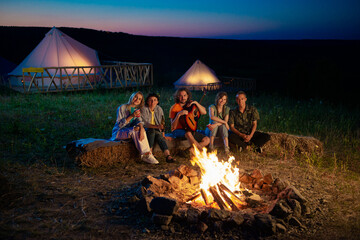 Charismatic and attractive happy group of friends have a good time at the campsite they singing playing on the guitar while sitting down on the haystack beside the fire stake