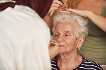 In this heartwarming real-life scene, a girl in a hijab and her sister lovingly apply makeup to their elderly grandmother, preparing her for a special family anniversary celebration, showcasing the