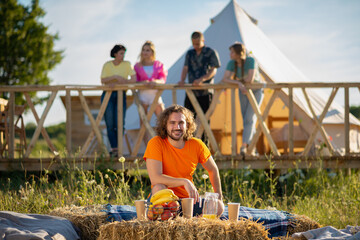 Posing in front of the camera good looking man smiling large while sitting down on the haystack at the campsite at the nature