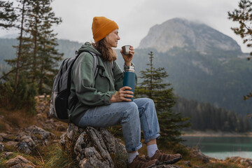 Young cheerful woman traveler with a backpack and thermos on the shore of a lake in the mountains. Solo travel concept - obrazy, fototapety, plakaty