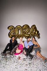 smiling multiethnic friends sitting near confetti and balloons with 2024 numbers, Happy New Year
