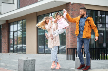 Fototapeta premium Full length of bearded man and blonde woman holding little girl and smiling while walking down the street in new urban district. Happy loving family with kid having fun outdoors.