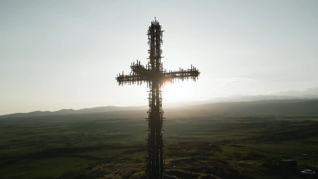 The largest iron cross in Armenia in the rays of the setting sun. On the background of high mountain Aragats in clear weather. Drone video. Middle plan, the camera is moving in an arc.