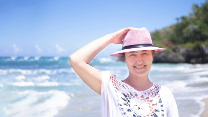 An elegant tourist in a white summer dress and a pink hat walks along the beach on a sunny day on the ocean. He looks into the camera and at the ocean, resting on the shore of a tropical beach.