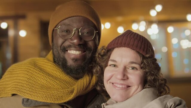 Portrait medium close-up shot of ethnically diverse family couple wearing warm clothes standing against cottage, looking at camera