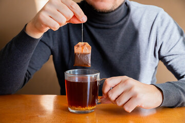 Male man holding wet teabag, brewing black vitamin tea with bag in transparent cup on table. Tea...
