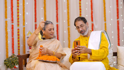 South-Indian couple preparing for the celebration of Diwali festival at home - using mobile for online shopping. Loving couple enjoying the festival of Diwali - making flower garlands at home  scro...