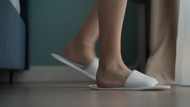 Woman puts on white slippers, getting out of bed on the floor in a hotel room. The guest puts her feet in slippers gets out of bed and leaves close-up. 