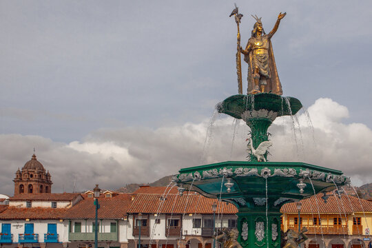 CUSCO, PERU - APRIL 30, 2022 : The statue dedicated to Inca emperor Manco Capac which sits above the water fountain at Plaza de Armas at Cusco in Peru. On left stands Church of the Society of Jesus.