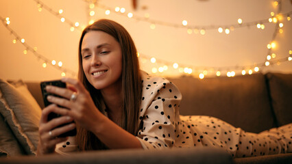Happy woman using smartphone in the evening at home while laying on sofa