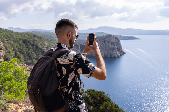 Caucasian man with short hair on a viewpoint taking a picture of the sea with his mobile phone. High angle view