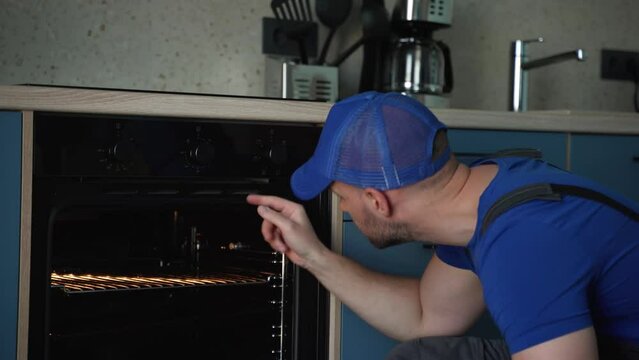 A man in a blue cap repairs an oven. Young Male Technician Repairing Oven In Kitchen.