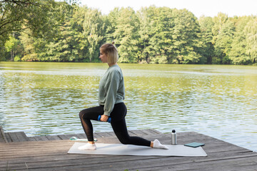 Fototapeta na wymiar Woman doing exercises in nature near the lake The girl is engaged in fitness. woman practicing yoga. The girl performs asanas. Yoga mat, bottled drinking water and stretch elastic band.