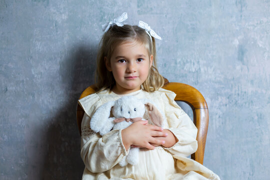 Horizontal portrait of adorable blond little girl in smart dress sitting on chair with toy bunny in her arms