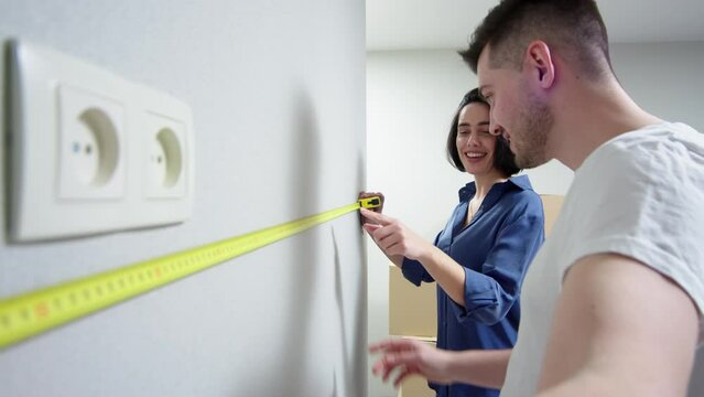 Video of a couple taking measurements on a wall. A young couple is discussing apartment renovation. Happy man and woman in love using measuring equipment measure the wall.